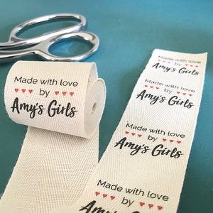 Op de afbeelding: Een rol wit katoenen lint met de tekst "Made with love by Amy's Girls" in zwart met rode hartjes gedrukt.