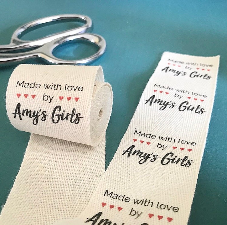 Op de afbeelding: Een rol wit katoenen lint met de tekst "Made with love by Amy's Girls" in zwart met rode hartjes gedrukt.