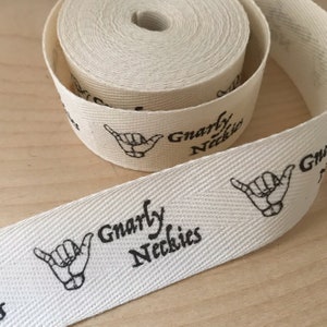 K&ouml;nnte beinhalten: Wei&szlig;es Baumwollband mit einem schwarzen Aufdruck einer Handgeste und dem Text "Gnarly Neckies".