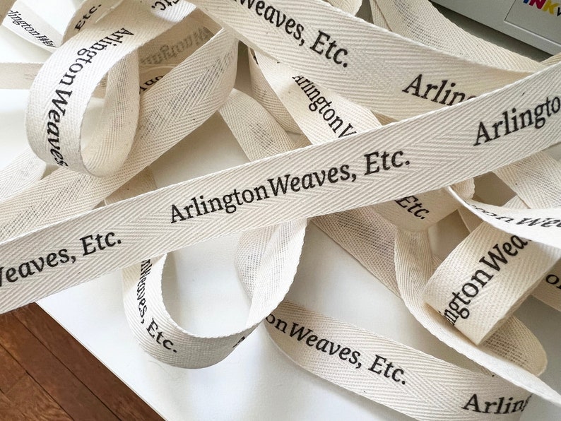 K&ouml;nnte beinhalten: Nahaufnahme von mehreren beigefarbenen Baumwollbandspulen mit dem aufgedruckten Text "Arlington Weaves, Etc.".