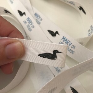 K&ouml;nnte beinhalten: Wei&szlig;es Stoffband mit einem schwarzen Loon-Vogel und dem Text "Mary on Lake Laski" darauf gedruckt.