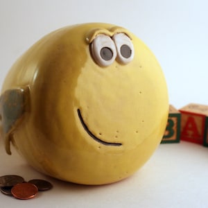 May include: A yellow ceramic piggy bank shaped like a bird with large white eyes and a black smile. The piggy bank is sitting on a white surface with wooden blocks that spell out "BABY" in front of it.