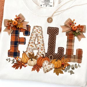 May include: A cream-coloured t-shirt featuring the word FALL in large, fabric-appliqued letters with varied patterns including plaid, floral, and polka dots. The design is accented with fabric pumpkins, leaves, and striped bows.