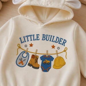 Puede incluir: Una sudadera con capucha de color crema que presenta orejas de animal bordadas en la capucha. El frente muestra el texto LITTLE BUILDER sobre un gráfico de tendedero con un babero, bota de trabajo, body azul y casco amarillo.