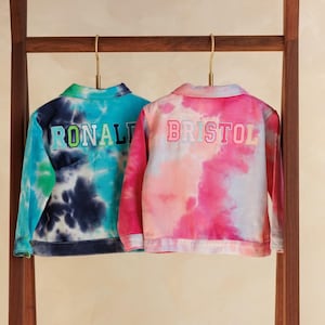 May include: Two tie-dye jackets hang on a wooden rack. One is blue and green with "RONALD" embroidered on the back. The other is pink and white with "BRISTOL" embroidered on the back. The jackets have long sleeves and a collared neckline.