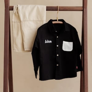 Puede incluir: Camisa negra con bolsillo blanco y el nombre "Adam" bordado en el pecho. Pantalones beige doblados y colgados en un estante de madera. La camisa tiene mangas largas y una pequeña etiqueta roja.