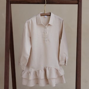 May include: A beige linen dress with long sleeves and a ruffled hem, hanging on a wooden clothing rack. The dress features a collared neckline and a button-down front. The rack is made of dark wood.