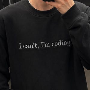 May include: Black sweatshirt with the text "I can't, I'm coding" in white. The person is holding a silver laptop with an Apple logo.