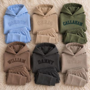 May include: Six matching hoodie and sweatpant sets in light blue, beige, olive green, brown, charcoal, and tan. Each hoodie features a hood, long sleeves, and a name embroidered in a collegiate font: GORDON, CHRIS, CALLAHAN, WILLIAM, DANNY, and RYAN.
