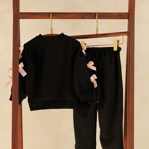 May include: A black two-piece outfit for children, consisting of a long-sleeved sweatshirt and matching pants. The sweatshirt and pants are adorned with pink bows on the sleeves and sides. The clothing set is hanging on a wooden rack.