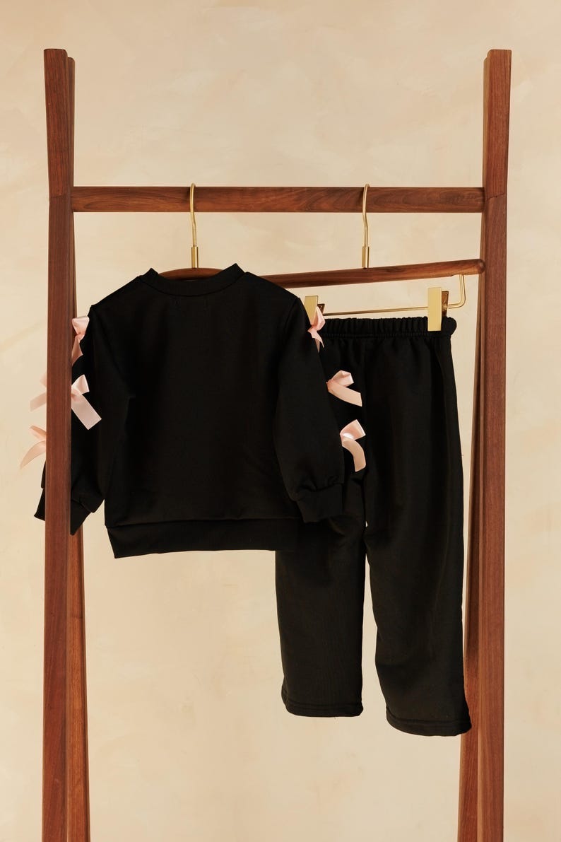 May include: A black two-piece outfit for children, consisting of a long-sleeved sweatshirt and matching pants. The sweatshirt and pants are adorned with pink bows on the sleeves and sides. The clothing set is hanging on a wooden rack.