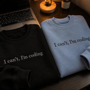 K&ouml;nnte beinhalten: Zwei Sweatshirts mit Rundhalsausschnitt, eines in Schwarz und eines in Hellblau, werden auf einer dunklen Holzoberfl&auml;che gezeigt. Jedes Sweatshirt hat den gestickten Text "I can't, I'm coding" in Wei&szlig;. Ein Laptop und eine kleine Lampe sind im Hintergrund zu sehen.