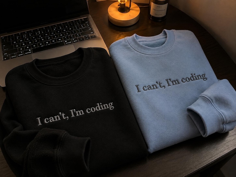 K&ouml;nnte beinhalten: Zwei Sweatshirts mit Rundhalsausschnitt, eines in Schwarz und eines in Hellblau, werden auf einer dunklen Holzoberfl&auml;che gezeigt. Jedes Sweatshirt hat den gestickten Text "I can't, I'm coding" in Wei&szlig;. Ein Laptop und eine kleine Lampe sind im Hintergrund zu sehen.