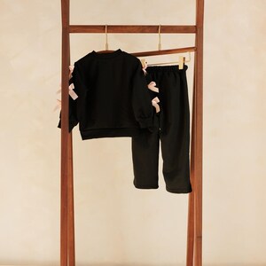 May include: A black two-piece outfit for children, consisting of a long-sleeved sweatshirt and pants, hangs on a wooden clothing rack. The sweatshirt and pants are adorned with pink bows. The rack is made of dark wood.