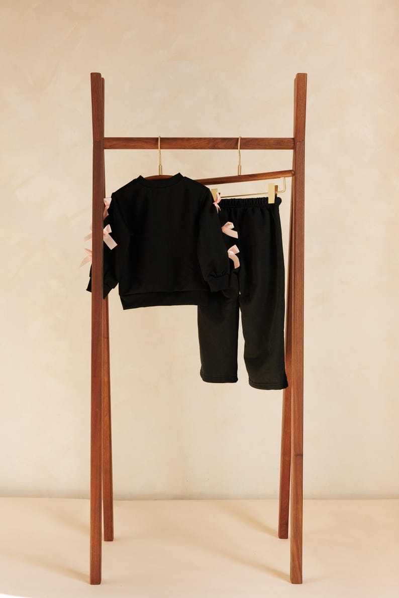 May include: A black two-piece outfit for children, consisting of a long-sleeved sweatshirt and pants, hangs on a wooden clothing rack. The sweatshirt and pants are adorned with pink bows. The rack is made of dark wood.
