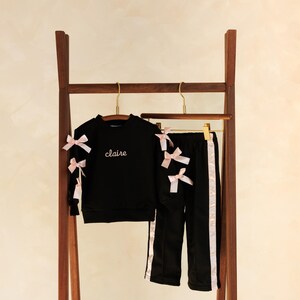 May include: Black children's tracksuit set with pink bows and trim. The set includes a sweatshirt with the name "claire" embroidered on the front and matching pants. The outfit is displayed on a wooden clothing rack.