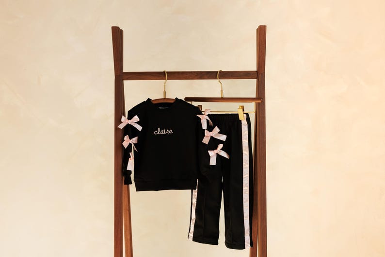 May include: Black children's tracksuit set with pink bows and trim. The set includes a sweatshirt with the name "claire" embroidered on the front and matching pants. The outfit is displayed on a wooden clothing rack.
