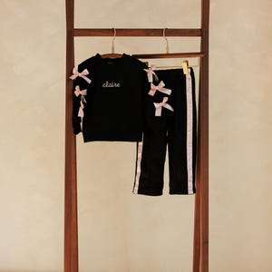 May include: A black two-piece outfit for children, including a long-sleeved top and pants, displayed on a wooden clothing rack. The top features the name "claire" embroidered in white, and both pieces are adorned with pink bows. The pants have pink stripes.