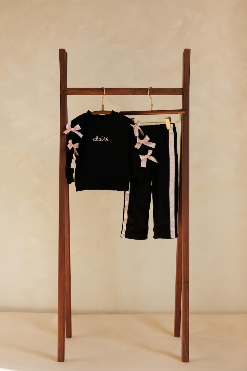 May include: A black two-piece outfit for children, including a long-sleeved top and pants, displayed on a wooden clothing rack. The top features the name "claire" embroidered in white, and both pieces are adorned with pink bows. The pants have pink stripes.