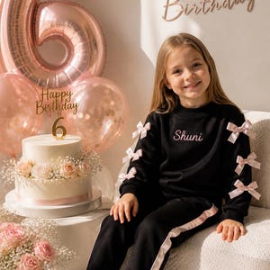 May include: A young person in a black sweatsuit with pink bow accents and a matching stripe on the pants. The word "Shuni" is embroidered on the sweatshirt. A birthday cake with a "6" candle and balloons are in the background.