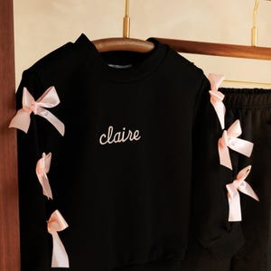 May include: Black sweatshirt and pants set with pink bow accents. The black sweatshirt has the name "claire" embroidered in white script. The sleeves of the sweatshirt are adorned with pink bows. The pants have a pink stripe down the side.