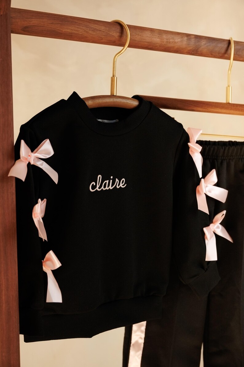 May include: Black sweatshirt and pants set with pink bow accents. The black sweatshirt has the name "claire" embroidered in white script. The sleeves of the sweatshirt are adorned with pink bows. The pants have a pink stripe down the side.
