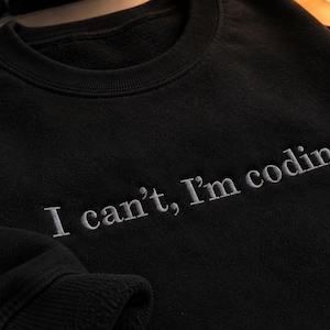 K&ouml;nnte beinhalten: Schwarzes Sweatshirt mit dem gestickten Text "I can't, I'm coding" in Hellgrau. Das Sweatshirt hat einen Rundhalsausschnitt und lange &Auml;rmel. Der Text ist mittig auf der Brust platziert.