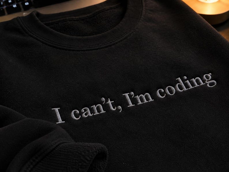 K&ouml;nnte beinhalten: Schwarzes Sweatshirt mit dem gestickten Text "I can't, I'm coding" in Hellgrau. Das Sweatshirt hat einen Rundhalsausschnitt und lange &Auml;rmel. Der Text ist mittig auf der Brust platziert.