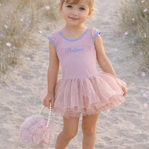 Może przedstawiać: Mała dziewczynka w różowym trykocie baletowym z warstwową tiulową spódnicą i imieniem "Polina" w skrypcie. Trzyma puszystą różową torebkę z perłową rączką. Strój jest ozdobiony jasnoniebieskimi wykończeniami.