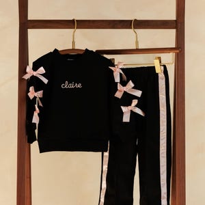 May include: Black children's outfit with pink bow accents. The set includes a sweatshirt with the name "claire" embroidered in cursive and matching pants with pink side stripes. The outfit is displayed on a wooden clothing rack.