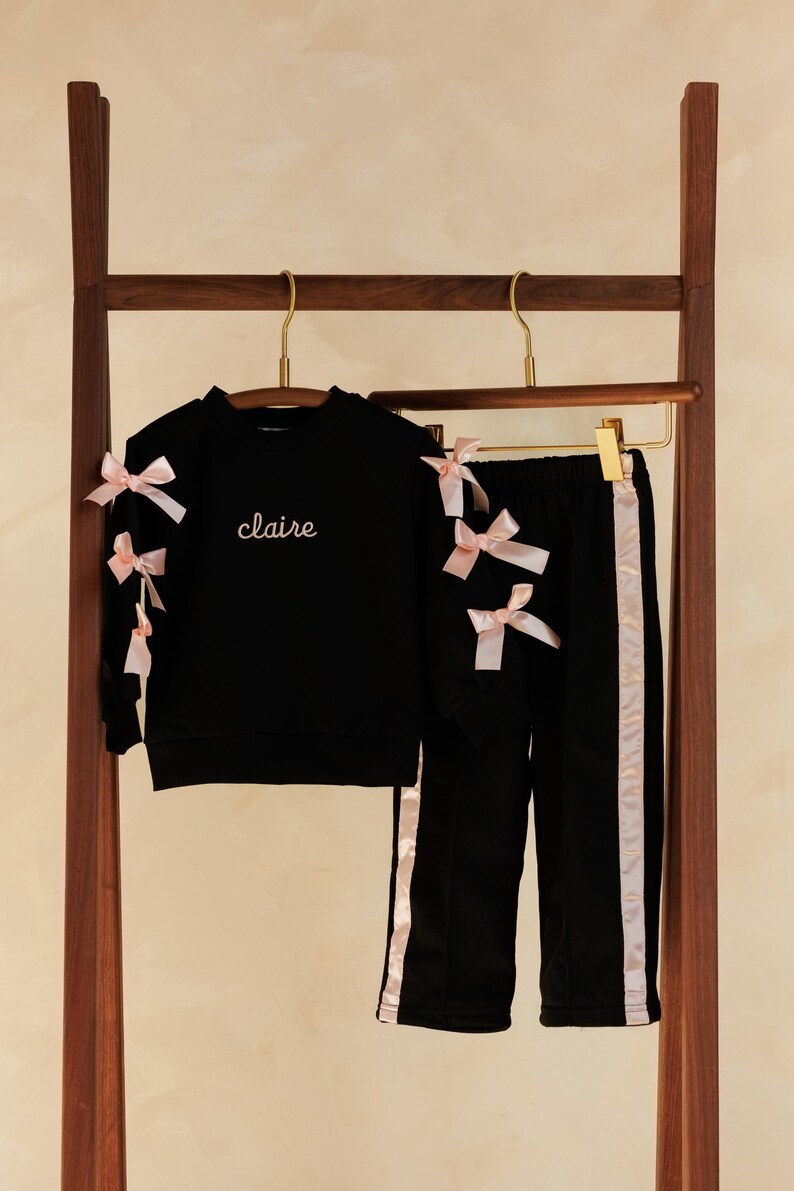 May include: Black children's outfit with pink bow accents. The set includes a sweatshirt with the name "claire" embroidered in cursive and matching pants with pink side stripes. The outfit is displayed on a wooden clothing rack.