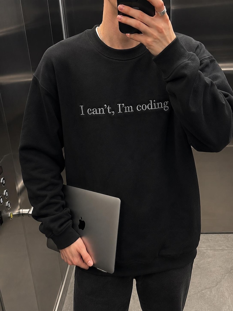 K&ouml;nnte beinhalten: Schwarzes Sweatshirt mit dem wei&szlig;en Text "I can't, I'm coding". Die Person h&auml;lt einen silbernen Laptop mit einem Apple-Logo.