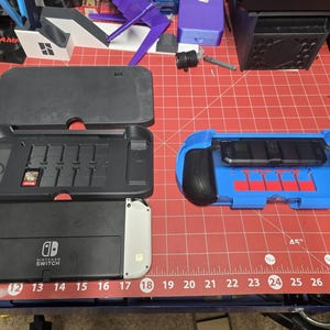 May include: A black and white Nintendo Switch console with a black case and a blue controller. The case has slots for game cartridges. The controller is blue with black grips and red accents. The text "SWITCH" is visible.