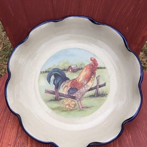 May include: A ceramic serving platter with a scalloped edge and a blue rim. The center features a painting of a rooster, a chick, and a fence in a rural setting. The plate is off-white with a glossy finish.