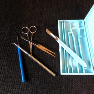 May include: A set of metal precision tools including a scalpel with a light blue handle, a pair of silver tweezers, a pair of silver scissors, and a thin metal probe with a blue handle, all arranged next to an open light blue plastic storage case.