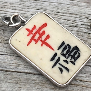 May include: A rectangular pendant with a silver-colored frame. The pendant features a cream-colored background with red and black Chinese characters. A silver loop is attached at the top for hanging.