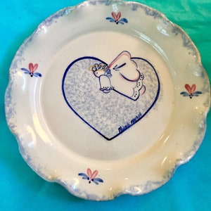 May include: White ceramic plate with a scalloped edge and a blue and pink floral design. The plate features a blue heart with a hand-drawn angel illustration. The plate is signed "Miranda 1995".