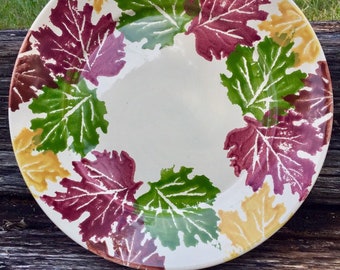 May include: A round, off-white ceramic plate with a leaf design. The leaves are in shades of burgundy, green, and yellow, arranged around the rim. The plate is set on a weathered wooden surface, suggesting a rustic or autumnal theme.