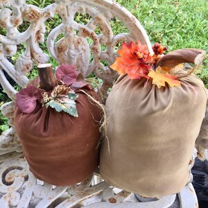 May include: Two fabric pumpkins in shades of brown and tan, decorated with faux fall leaves and stems. The pumpkins are sitting on a white, ornate bench, creating a rustic autumn theme.