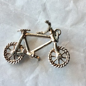 May include: A small, silver-colored bicycle charm. The bicycle has detailed features, including a seat, handlebars, and wheels with a rope-like design. The charm is likely made of metal and is designed to be a pendant or charm.