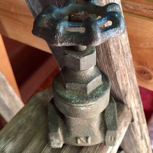 May include: A vintage bronze water valve with a star-shaped handle. The handle is painted blue and shows signs of wear. The valve is mounted on a wooden surface, showcasing its aged appearance and industrial design.