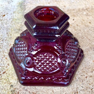 May include: A deep red, hexagonal glass candle holder. The candle holder has a textured, patterned design with a geometric pattern and a scalloped edge. The top section is also hexagonal and holds the candle.