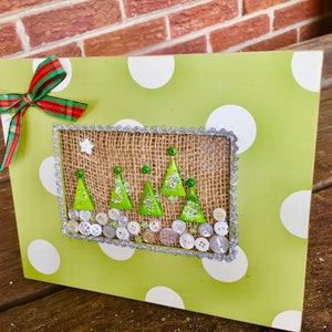 May include: A Christmas-themed decorative wooden plaque. The plaque features a light green background with white polka dots. A burlap-framed scene depicts green Christmas trees, buttons, and a star. A red and green plaid bow adorns the top.