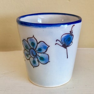May include: A small, white ceramic cup with a blue rim and floral designs. The cup features hand-painted blue flowers with black outlines. The design includes a large flower and a smaller, stylized flower. The cup is approximately 3 inches tall.