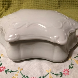 May include: A white porcelain trinket box with a lid. The box has a scalloped edge and decorative embossed detailing. The box is sitting on a white cloth with floral embroidery.