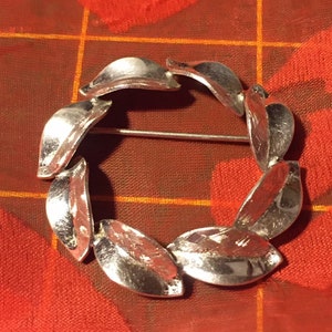 May include: A silver brooch in the shape of a wreath of leaves. The leaves are textured and have a slightly hammered look.