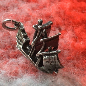 May include: A silver charm in the shape of a sailing ship with a raised, detailed design. The ship has a single mast and a sail with a decorative pattern. The charm has a loop at the top for attaching to a bracelet or necklace.