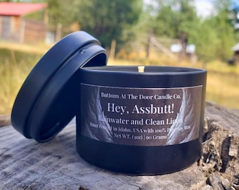 May include: A black candle tin with a lid, resting on a wooden surface. The label reads "Hey, Assbutt! Rainwater and Clean Linen." The candle is hand-poured in Idaho, USA, and contains 90 grams of soy wax.