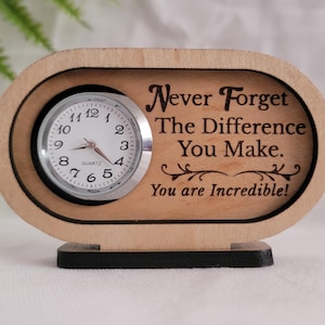 May include: An oval-shaped light wood desk clock with a black base. The clock face is white with black numbers and hands. The wood is engraved with the text: Never Forget The Difference You Make. You are Incredible! with decorative flourishes.
