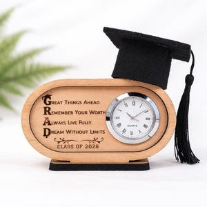 May include: A wooden graduation clock with a black mortarboard hat and tassel. The clock face is white with black Roman numerals. The wooden base has the word "GRAD" spelled out vertically with inspirational phrases and "CLASS OF 2026" below.